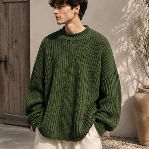 Puede incluir: Un suéter de punto grueso en color verde bosque con textura acanalada, cuello alto y mangas largas con hombros caídos. La prenda se combina con pantalones de color beige claro, creando un conjunto sencillo y superpuesto con un ajuste relajado.