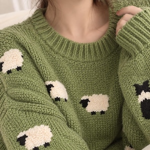 Puede incluir: Un suéter de color verde salvia con cuello redondo que presenta un patrón repetitivo de pequeñas ovejas blancas texturizadas con caras y patas negras. La prenda de punto tiene un cuello acanalado y mangas largas de tejido grueso.