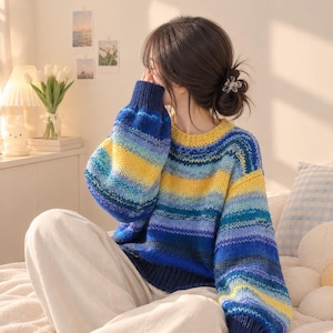 May include: A long-sleeved knit jumper featuring a horizontal striped pattern in shades of yellow, light blue, medium blue, and dark blue. The garment has a round neckline and ribbed cuffs and hem in a solid dark blue colour.