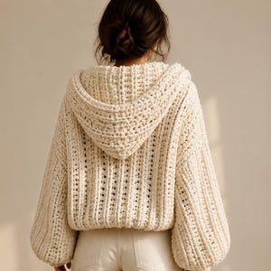 May include: A cream-colored, chunky knit hooded sweater featuring a thick, vertical ribbed texture, long sleeves with dropped shoulders, and a relaxed, cropped fit. The garment is crafted from a heavy, soft yarn and includes a hood.