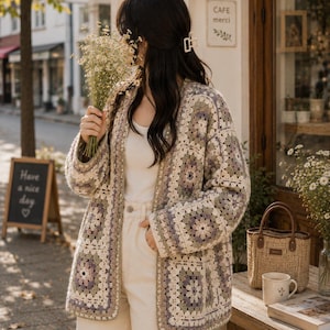 May include: A long-sleeved cardigan made of crocheted granny squares in shades of cream, sage green, and muted purple. The garment features an open front, a straight hem, and a relaxed fit, worn over a plain white top and light-coloured trousers.