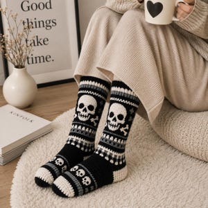 May include: A pair of black, white, and gray mid-calf socks featuring a repeating skull and crossbones pattern. The knit design includes horizontal stripes and small skull motifs near the toe area, crafted with a textured, thick yarn construction.
