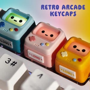 Könnte beinhalten: Drei farbenfrohe Retro-Arcade-Tastenkappen in Blau, Rosa und Gelb, jede mit einem kleinen Bildschirm und einem niedlichen Gesicht. Die Tastenkappen befinden sich auf einer weißen Tastatur, mit dem Text "RETRO ARCADE KEYCAPS" in Lila.