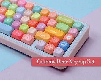 May include: A colorful keyboard with pastel-colored keycaps. The keys are in shades of pink, yellow, green, blue, and orange. The keyboard has a light blue frame and the text "Gummy Bear Keycap Set" is displayed on a red banner.