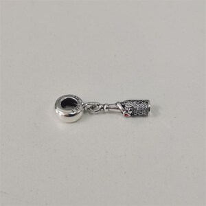 Puede incluir: Un charm de plata con un diseño de botella. El charm presenta una botella con una superficie texturizada y un pequeño detalle de corazón rojo. El charm está conectado a un lazo redondo con el texto "S 925".