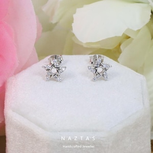 May include: A pair of silver star-shaped stud earrings with small, clear gemstones. The earrings are displayed on a white surface with a soft-focus floral background. The brand name "NAZTAS" and the tagline "Handcrafted Jeweller" are visible.