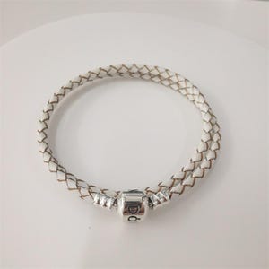 May include: A double-wrap bracelet featuring a white braided leather cord. The bracelet includes a cylindrical silver-toned metal clasp at the centre, which is engraved with the text PANDORA and a small crown symbol above the letter O.