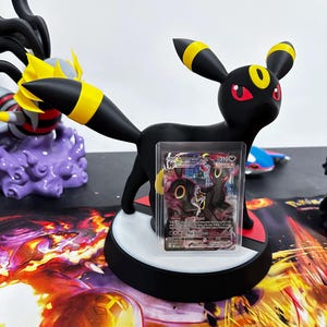 Puede incluir: Una figura negra de Umbreon con detalles amarillos y ojos rojos, sobre una base circular. Una carta de Pokémon se muestra frente a la figura. La base es negra con un diseño circular blanco y rojo. El fondo presenta una superficie colorida y estampada.