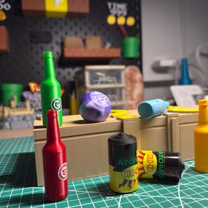 May include: Miniature novelty items including red, green, and yellow bottles, a purple sphere, and a black and yellow battery. The items are arranged on a green grid surface, with a black pegboard background.