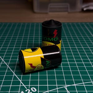 May include: Two black and yellow film canisters with the text "COMO" and "AT". One canister is upright, the other is lying on its side. The canisters are on a green cutting mat with white grid lines and the number 120.