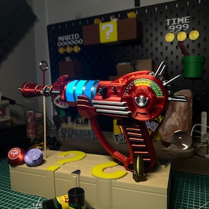 Peut inclure: Un pistolet laser rouge et argenté avec des lumières bleues, une poignée et une jauge. Le pistolet est posé sur une plateforme en bois avec des découpes en forme de point d'interrogation et deux piles cylindriques. Un arrière-plan sur le thème de Mario est visible.