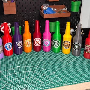 May include: A display of brightly coloured bottles, each with a unique logo. The bottles are arranged on a green cutting mat. The bottles are in various colours, including black, blue, brown, purple, red, pink, green, yellow, and grey.