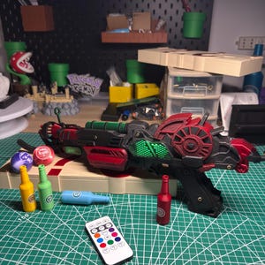 以下が含まれることがあります： 未来的なデザインの、赤、黒、緑色の詳細な小道具銃。カラフルなボトルとリモコンが付属しています。背景には、コインと疑問符ブロックを備えたマリオをテーマにしたディスプレイがあります。小道具銃の長さは約38cmです。