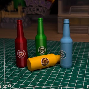 Op de afbeelding: Vier kleurrijke, flesvormige objecten. Drie staan rechtop: rood, groen en blauw. Een gele fles ligt op zijn kant. Elk heeft een cirkelvormig embleem. Op een groene rasterondergrond.