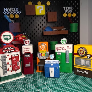 Può includere: Una collezione di sei distributori di potenziamenti per videogiochi in miniatura, tra cui Mule Kick, Juggernog, Stamin-Up, Quick Revive, Speed Cola e Double Tap. Questi colorati modelli rettangolari in plastica presentano loghi, gettoniere e pulsanti dettagliati, esposti su un tappetino da taglio verde davanti a un pannello forato nero con decorazioni murali a tema Mario.