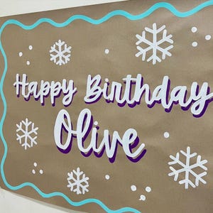 May include: A brown paper birthday banner with the words "Happy Birthday Olive" in white and purple lettering. The banner is decorated with white snowflakes and a teal border.