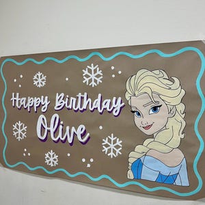 May include: A brown banner with a turquoise border and the words "Happy Birthday Olive" in white and purple. The banner features a cartoon image of Elsa from Frozen and snowflake designs.