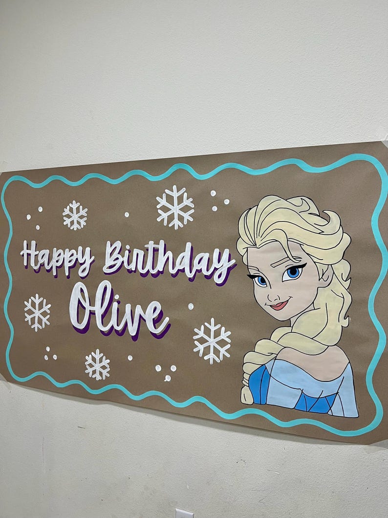 May include: A brown banner with a turquoise border and the words "Happy Birthday Olive" in white and purple. The banner features a cartoon image of Elsa from Frozen and snowflake designs.