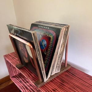 May include: A wooden record holder with copper pipe accents, holding several vinyl records. The holder sits on a red-stained wooden shelf. The records are visible, with the word "JOURNEY" on one album cover.