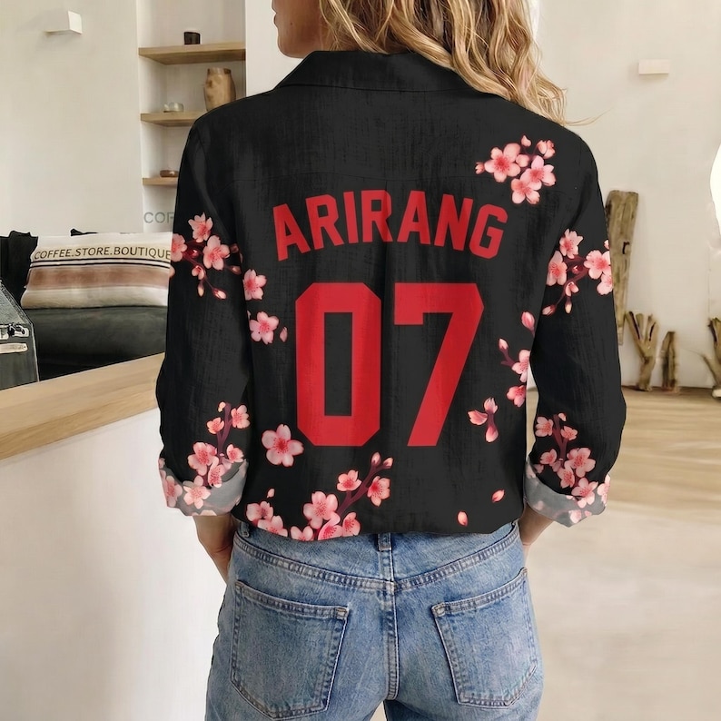 May include: A black long-sleeve button-down shirt featuring a red printed graphic that reads ARIRANG above the number 07. The shirt is decorated with a pattern of pink cherry blossom flowers across the back and sleeves, paired with blue jeans.