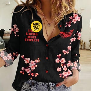 May include: A black long-sleeve button-down shirt featuring a pink cherry blossom floral pattern, a red graphic of a whale, and red text that reads "BTS WORLD TOUR ARIRANG". The shirt has a V-neckline and is paired with blue denim jeans.