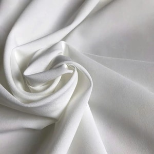 May include: A solid white fabric with a smooth, matte texture, draped in soft, flowing folds that create deep shadows and highlights. The material appears opaque and consistent in color, forming a swirling, circular pattern in the center.