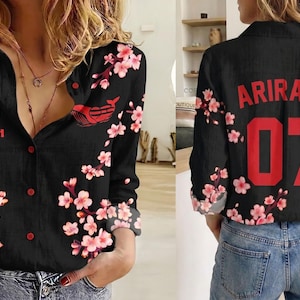 May include: A black long-sleeve button-down shirt featuring a pink cherry blossom floral pattern. The front displays the text "ITS WORLD TOUR ARIRANG" and a small red graphic. The back features the red text "ARIRANG" above the large red number "07".
