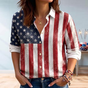 May include: Button-down shirt featuring the American flag design. The left side of the shirt displays a blue field with white stars, while the right side has red and white stripes. The shirt has long sleeves and a white collar and cuffs.