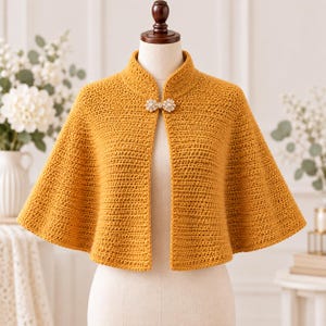 Può includere: Una mantellina all'uncinetto color giallo senape con collo alto e una chiusura decorativa a forma di fiocco impreziosita da piccole perle bianche. Il capo è esposto su un manichino e presenta una lavorazione a maglia testurizzata.