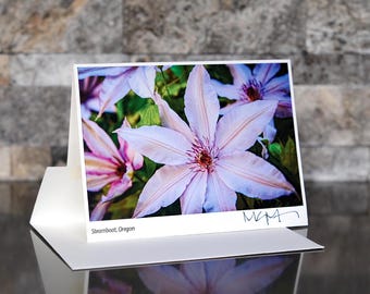 May include: A greeting card featuring a close-up photograph of a light pink clematis flower with a dark red center. The card is displayed open, with the text "Steamboat, Oregon" at the bottom. The card is on a white surface.