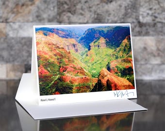 May include: A white greeting card featuring a vibrant landscape photograph of a canyon with red, green, and blue hues. The card is displayed in a white holder, with the text "Kauai, Hawaii" visible. Signed with a blue signature.