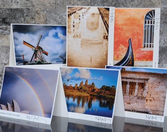 May include: A collection of six photographic postcards featuring travel destinations. Images include a windmill, a bridge, a building with a gondola, a rainbow over a building, a temple, and a building with statues. Each card has a white border.