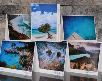 May include: A collection of six landscape greeting cards featuring scenic views of the ocean, beaches, and mountains. Each card displays a unique photograph with vibrant colors and natural elements. The cards are arranged in a semi-circular display.