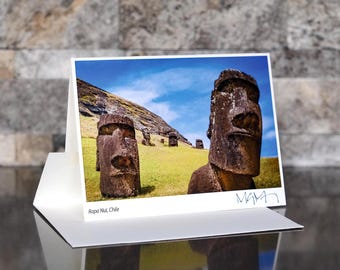 May include: A photograph of Easter Island's Moai statues on a greeting card. The image shows several large, weathered stone statues on a grassy hillside under a blue sky. The card is propped up on a white stand, with the text "Rapa Nui, Chile" visible.