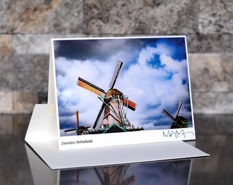 May include: A photograph of windmills in Zaanndam, Netherlands, displayed on a white card. The windmills have brown and orange blades against a cloudy blue sky. The card is propped up on a white stand, with the text "Zaanndam, Netherlands" at the bottom.