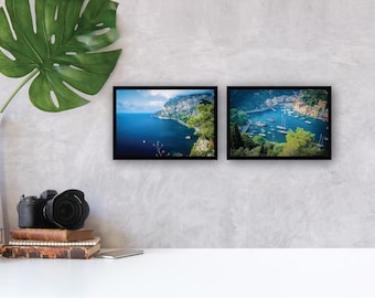 May include: Two framed landscape prints with black borders hang on a gray wall. One depicts a coastal view with blue water and cliffs. The other shows a harbor with boats. A camera, books, and a vase with a large leaf are on a white surface.