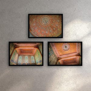 May include: Three framed prints displaying architectural details. The images show ornate ceilings with intricate patterns in shades of red, orange, and gold. The frames are black, and the prints are displayed against a textured grey wall.