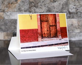 May include: A greeting card featuring a photograph of a weathered wooden door in San Miguel de Allende, Mexico. The door is framed by a yellow and red wall. The card is white and has the text "San Miguel de Allende, Mexico" and a signature.