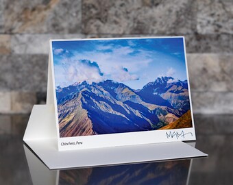 May include: A white greeting card featuring a scenic photograph of the Chinchero mountains in Peru. The image displays a range of blue and brown mountains under a cloudy sky. The card is displayed in a white holder with the text "Chinchero, Peru" and a signature.