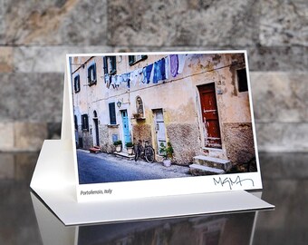 May include: A photograph of a street scene in Portofino, Italy, featuring aged buildings with colorful doors and windows. Laundry hangs overhead. A bicycle leans against a wall. The card is displayed in a white envelope.