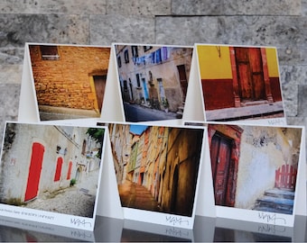 May include: A set of six greeting cards featuring photographs of European street scenes. The images showcase colorful buildings, doors, and alleyways. Each card is displayed on a white stand, with the artist's signature at the bottom.