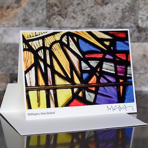 May include: A greeting card featuring a colorful stained-glass window design. The artwork includes yellow, blue, purple, and red hues, with black lines defining the shapes. The card is white, with the text "Wellington, New Zealand" and a signature.