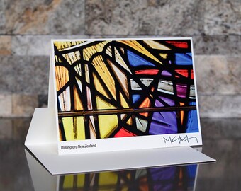 May include: A greeting card featuring a colorful stained-glass window design. The artwork includes yellow, blue, purple, and red hues, with black lines defining the shapes. The card is white, with the text "Wellington, New Zealand" and a signature.