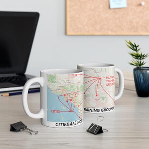 Puede incluir: Dos tazas de cerámica blanca con un diseño de mapa. Una taza muestra la palabra "CITIES" con flechas rojas que apuntan a "MILITARY TRAINING GROUNDS". El texto "CITIES ARE NOT" está en la parte inferior de la taza.