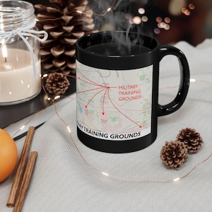 Puede incluir: Taza de cerámica negra con un diseño de mapa y flechas rojas que apuntan a "MILITARY TRAINING GROUNDS". La taza tiene un asa curva y el texto "MILITARY TRAINING GROUNDS" está impreso en la parte inferior. Sale vapor de la taza.