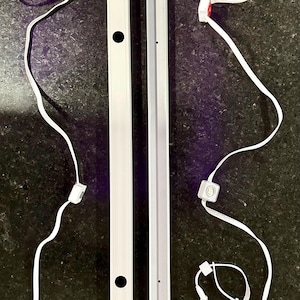 Puede incluir: Dos barras de luz LED blancas con diseño de canal y cableado blanco con pequeños conectores cuadrados. Una barra tiene un círculo negro, la otra un pequeño agujero. El fondo es una superficie oscura y texturizada.