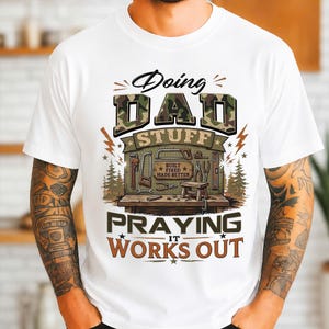May include: A white t-shirt featuring a graphic with the text Doing DAD STUFF PRAYING IT WORKS OUT. The design includes camouflage lettering, a workbench with various tools, a small stool, and a sign that reads BUILT FIXED MADE BETTER.