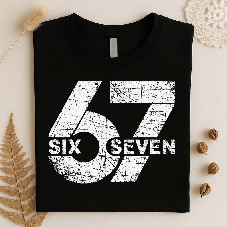 May include: A black crew neck t-shirt featuring a large, distressed white graphic of the number 67. The text SIX SEVEN is printed in a bold, white, weathered font across the center of the numbers, creating a vintage, worn-in aesthetic.