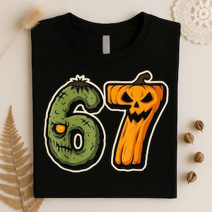 May include: A black t-shirt featuring a graphic of the number 67. The number 6 is designed as a green, textured zombie head with a single glowing yellow eye, while the number 7 is shaped like an orange jack-o'-lantern with a carved face.