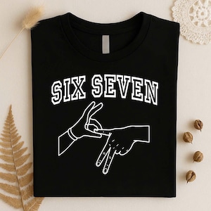 May include: A black t-shirt featuring the white text SIX SEVEN in a collegiate font arched above a white line-art graphic of two hands forming a hand sign. The design is centered on the chest of the shirt, which has a standard crew neckline.